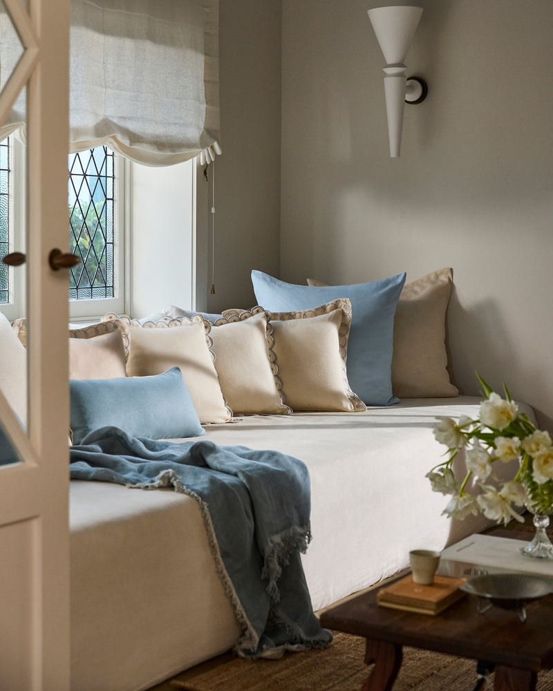 A cozy living room corner with a cream sofa, blue pillows, a Bed Threads Crème & Coast 100% French Flax Linen Swirl Cushion Cover, a light blue throw, wooden coffee table with flowers, and a window with sheer roman shade streaming in light.
