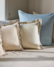 A close-up of a bed with two cream Bed Threads Crème & Coast 100% French Flax Linen Swirl Cushion Covers and one large light blue linen pillow, arranged against a beige wall. - view