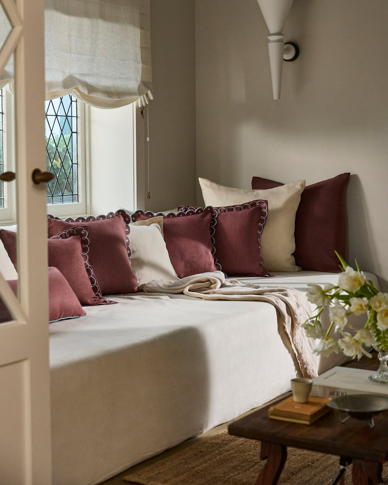 A cozy daybed with cream French flax linen bedding is styled with maroon and beige pillows near a patterned glass window. The inviting setting features the Bed Threads Cacao & Coast Swirl Cushion Cover, a soft throw, and a wooden table.