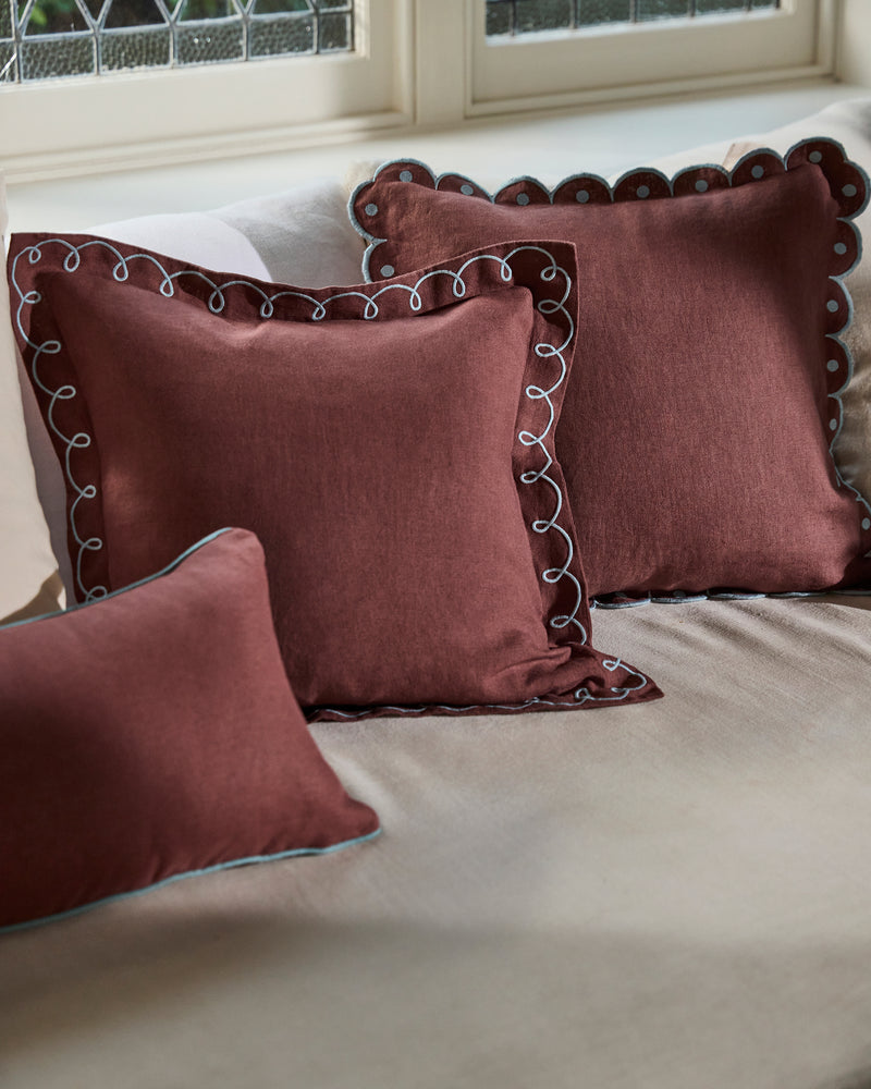 Three Cacao & Coast 100% French Flax Linen Swirl Cushion Covers by Bed Threads—two square, one rectangular with decorative white trim—are arranged on a beige sofa near a window with patterned glass.