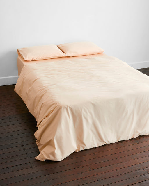 A minimal room features a neatly made bed with the Bed Threads Vanilla 100% Organic Cotton Duvet Cover Set, two pillows, and a dark wooden floor against a white wall.