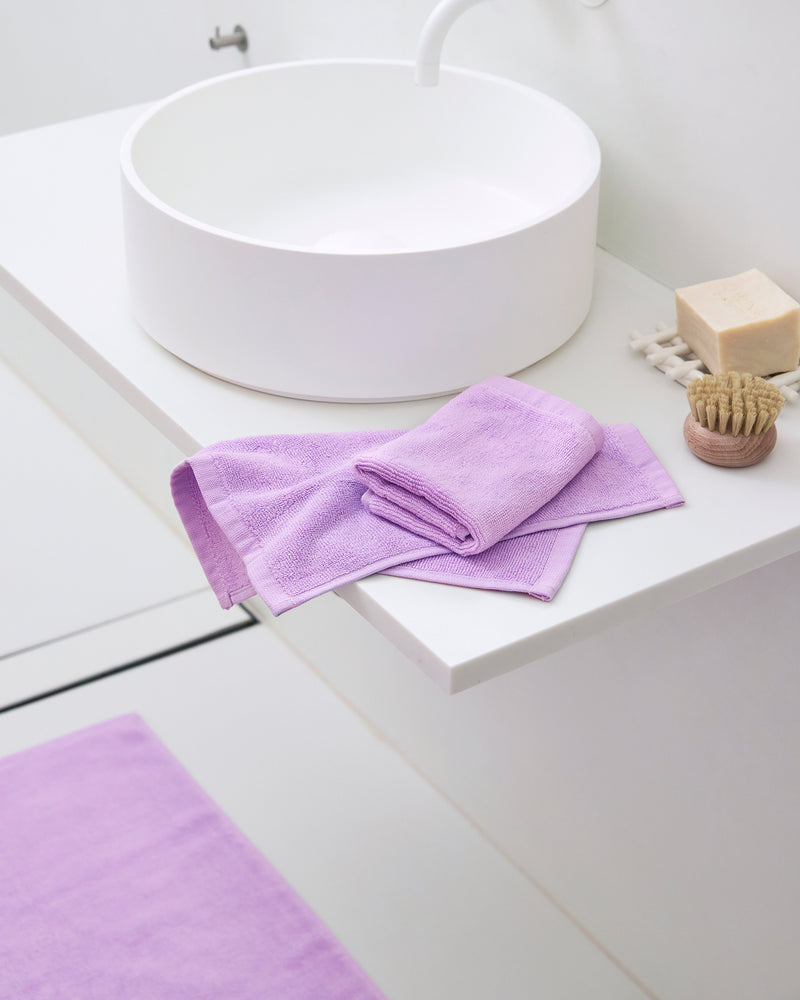 A modern white bathroom sink with a stack of Bed Threads’ Violet 100% Cotton Terry Face Washers (Set of Two), plus a beige soap bar, wooden brush, and a matching violet bath mat on the floor.