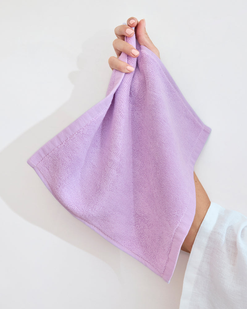 A hand displays a neatly folded Bed Threads Violet 100% Cotton Terry Face Washer (Set of Two), crafted from absorbent, skin-friendly fabric, against a plain white background.