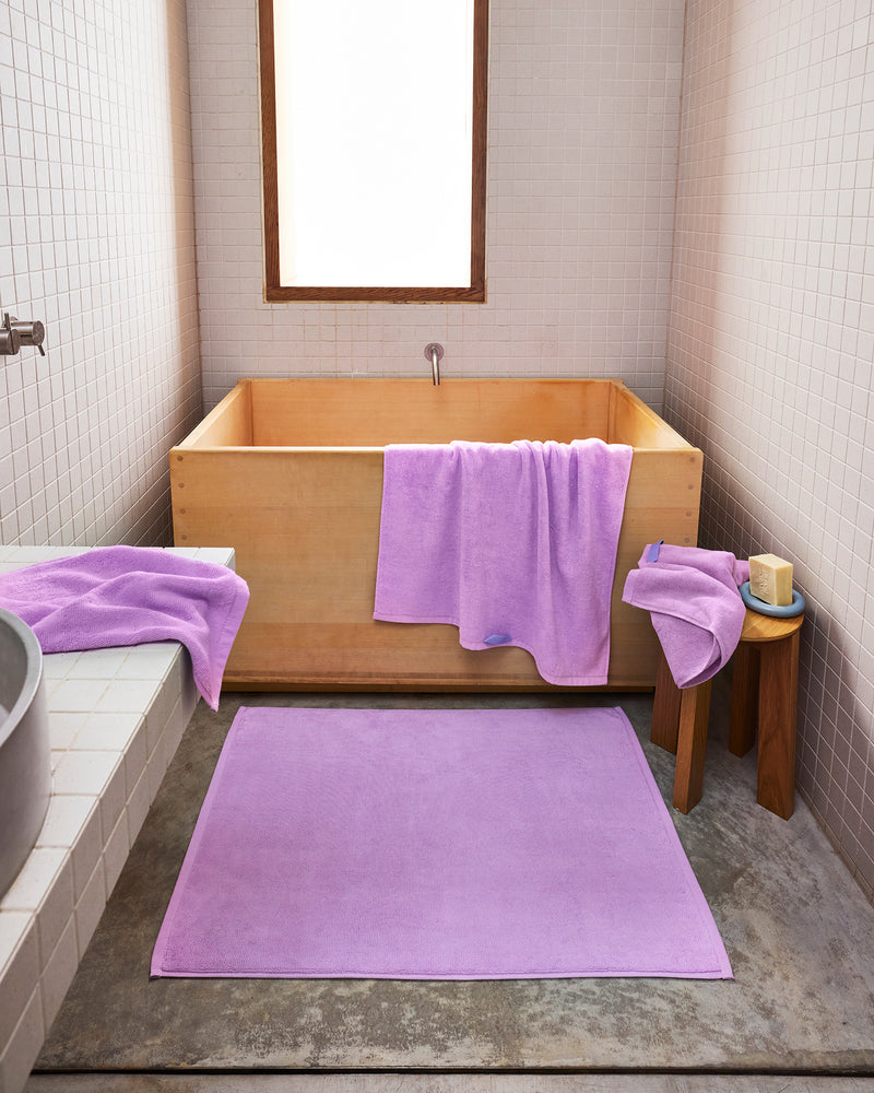 A modern bathroom features white tiled walls, a wooden soaking tub, and the Bed Threads Violet 100% Cotton Terry Bath Mat with light purple towels and washcloths. A stool beside the tub holds a towel, candle, and brush for added comfort.