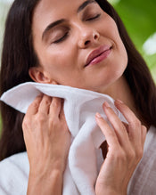 A woman with closed eyes gently smiles as she holds a Bed Threads Crisp White 100% Cotton Terry Face Washer (Set of Two) to her neck and face, evoking relaxation and self-care. Blurred greenery appears in the background. - view