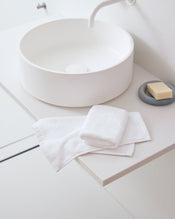 Crisp White 100% Cotton Terry Face Washers by Bed Threads, soft and absorbent, are folded beside a bar of soap on a gray dish atop a minimalist white bathroom sink with a round basin and curved faucet—gentle on skin. - view