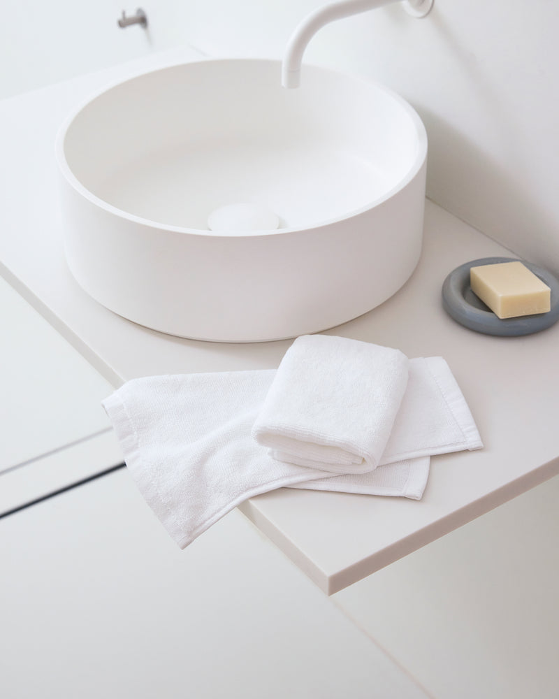 Crisp White 100% Cotton Terry Face Washers by Bed Threads, soft and absorbent, are folded beside a bar of soap on a gray dish atop a minimalist white bathroom sink with a round basin and curved faucet—gentle on skin.