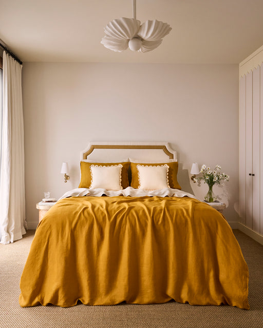 A cozy bedroom with a Bed Threads Turmeric 100% French Flax Linen Duvet Cover, white and yellow pillows, white bedside tables with lamps, a flower vase, modern ceiling light, and neutral-toned curtains and walls.