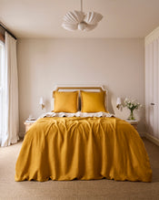 A neatly made bed features Bed Threads’ Turmeric & Crème 100% French Flax Linen Swirl European Pillowcases, paired with a mustard yellow comforter, against a beige wall. Two tables with lamps and flowers sit under a white pendant light. - view
