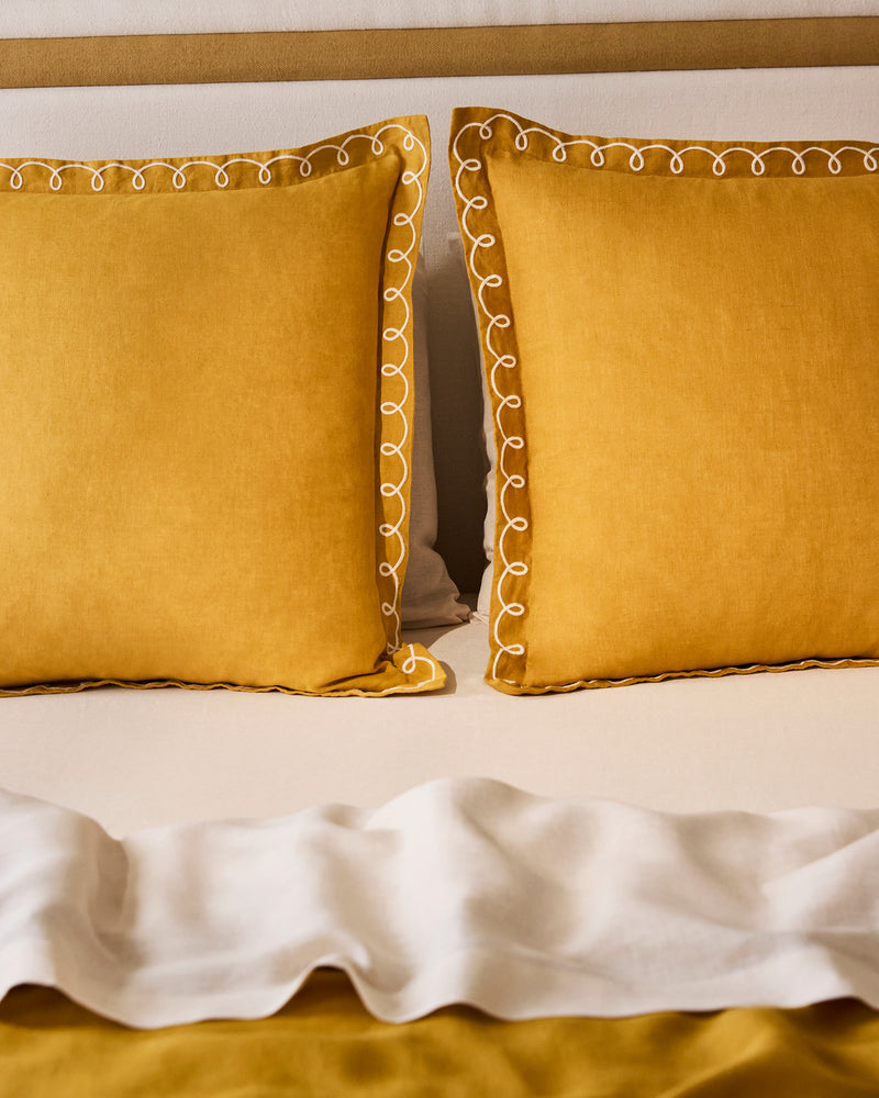 Two Bed Threads Turmeric & Crème 100% French Flax Linen Swirl European Pillowcases rest against a beige headboard. A white ruffled blanket is folded at the end, partly covering a 170GSM French flax linen mustard yellow bedspread.
