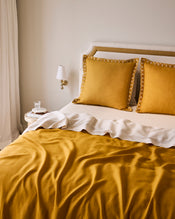 A neatly made bed features Bed Threads' Turmeric & Crème 100% French Flax Linen Swirl European Pillowcases, paired with a matching duvet and white throw. A round nightstand with a cup and saucer sits nearby, beneath a wall sconce. - view
