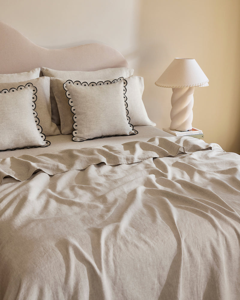 A neatly made bed features light beige French flax linen bedding with two Bed Threads Oatmeal & Charcoal 100% French Flax Linen Arch Cushion Covers, each with black scalloped edges, beside a sculptural white table lamp on the bedside table.