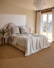A cozy, sunlit bedroom with a neatly made bed featuring Bed Threads Oatmeal & Charcoal 100% French Flax Linen Swirl European Pillowcases, decorative pillows, a wooden nightstand, a lamp, and French doors with sheer curtains to a balcony. - view