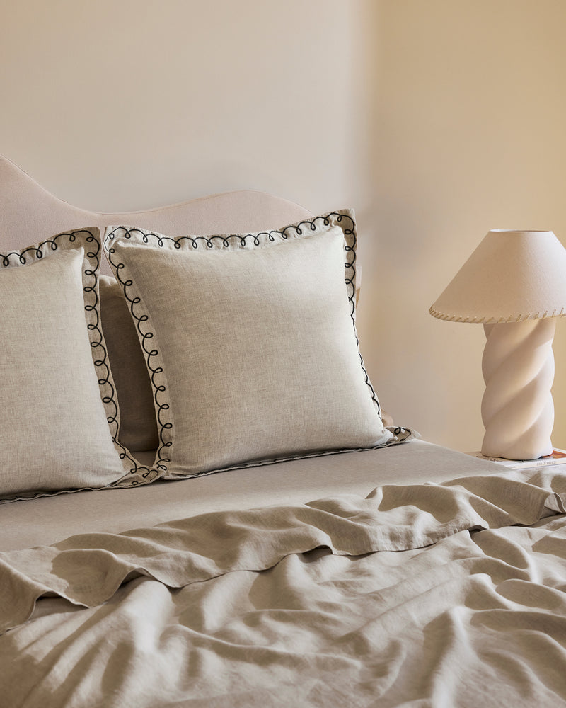 A neatly made bed with beige French flax linen and two Bed Threads Oatmeal & Charcoal 100% French Flax Linen Swirl European Pillowcases beside a small table topped with a cream spiral lamp and matching shade.