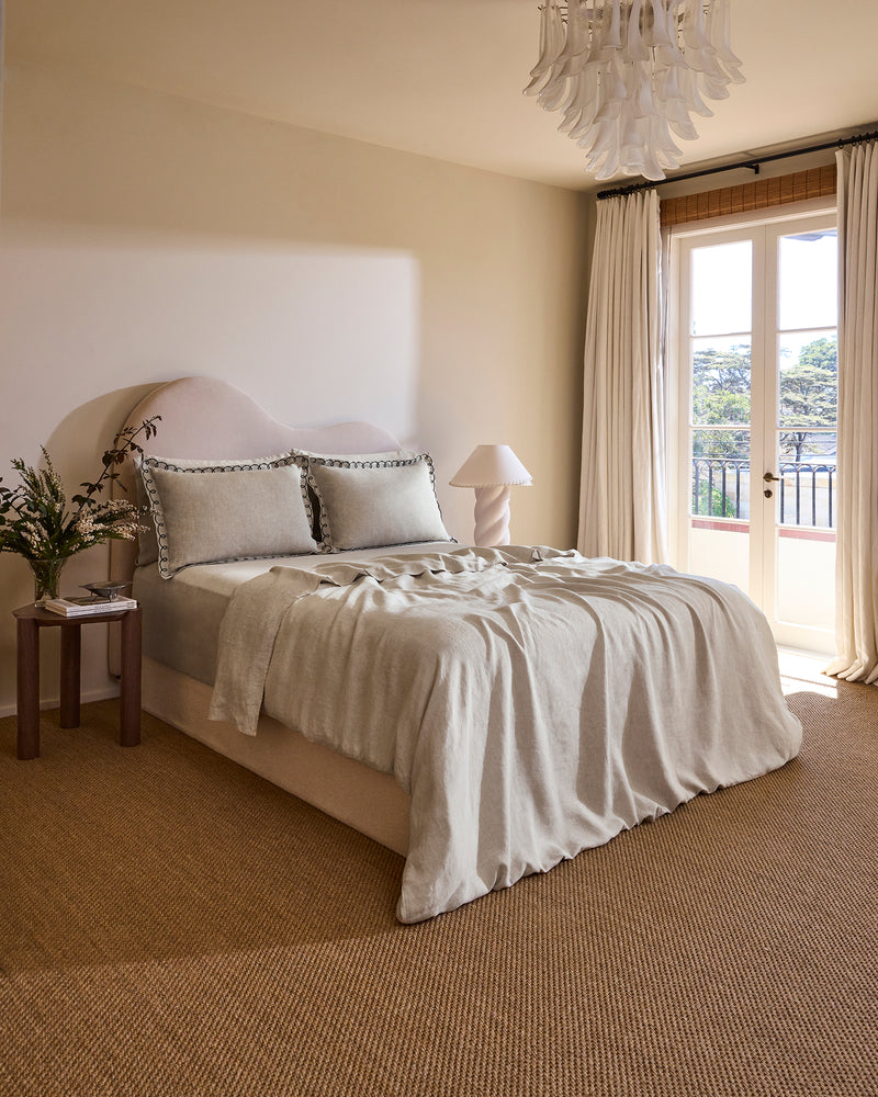 A cozy bedroom features a neatly made bed with Bed Threads’ Oatmeal & Charcoal 100% French Flax Linen Swirl Pillowcases, a small bedside table with a plant and lamp, and sheer curtains letting natural light in through a large window.