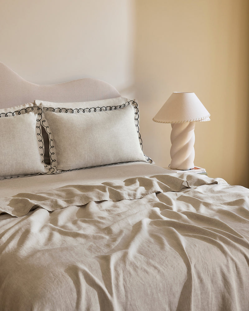 A neatly made bed with Bed Threads’ Oatmeal & Charcoal 100% French Flax Linen Swirl Pillowcases, set of two, sits in neutral tones beside a bedside table featuring a sculptural ceramic lamp and pleated lampshade.