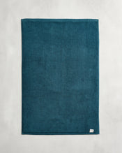The Bed Threads Aegean 100% Cotton Terry Bath Mat, in a teal blue rectangular design, is laid flat on a light gray surface with a small white tag visible at its bottom right corner. - view