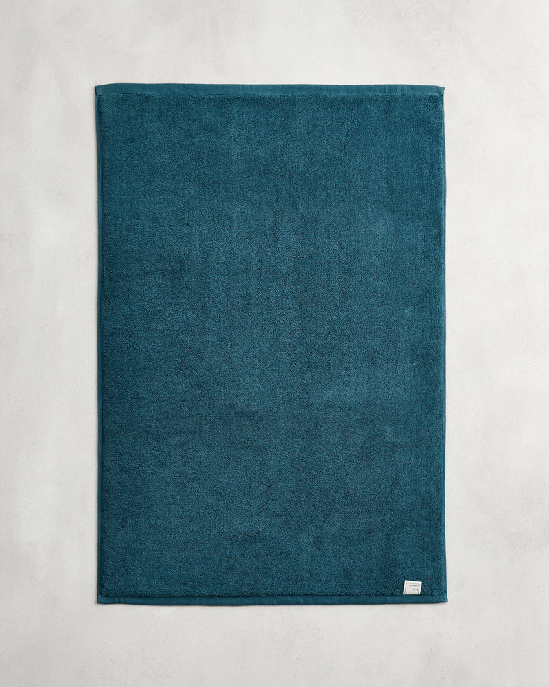 The Bed Threads Aegean 100% Cotton Terry Bath Mat, in a teal blue rectangular design, is laid flat on a light gray surface with a small white tag visible at its bottom right corner.