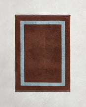 The Bed Threads Cacao & Lagoon Two-Toned 100% Cotton Terry Bath Mat features a rectangular brown design with a light blue border, shown here on a light gray textured surface. - view