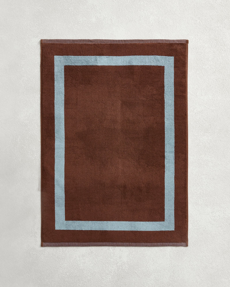 The Bed Threads Cacao & Lagoon Two-Toned 100% Cotton Terry Bath Mat features a rectangular brown design with a light blue border, shown here on a light gray textured surface.