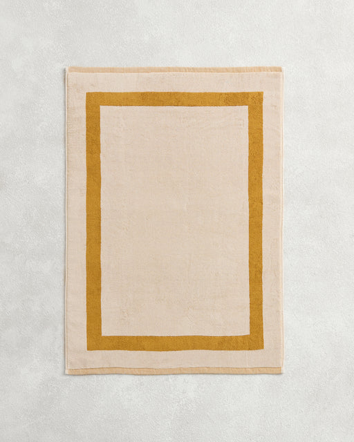 The Bed Threads Crème & Turmeric Two-Toned 100% Cotton Terry Bath Mat features a plush beige center with a bold mustard border, adding cozy style to your space.