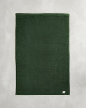 The Bed Threads Moss 100% Cotton Terry Bath Mat, in dark green, lies flat on a light gray surface with a small white tag visible in one corner. - view