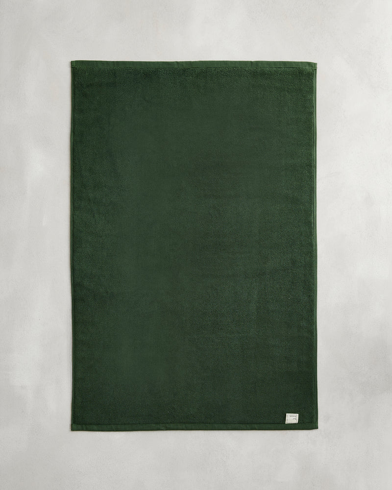 The Bed Threads Moss 100% Cotton Terry Bath Mat, in dark green, lies flat on a light gray surface with a small white tag visible in one corner.