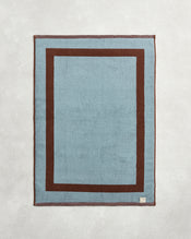 The Bed Threads Cacao & Lagoon Two-Toned 100% Cotton Terry Bath Mat features a rectangular light blue center, thick dark brown border, and a small fabric tag in one corner, set on a light textured surface. - view