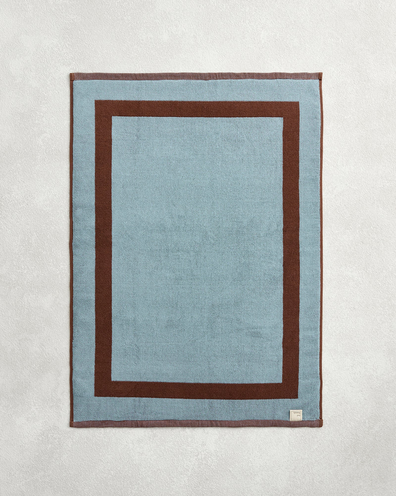 The Bed Threads Cacao & Lagoon Two-Toned 100% Cotton Terry Bath Mat features a rectangular light blue center, thick dark brown border, and a small fabric tag in one corner, set on a light textured surface.