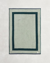 The Bed Threads Forest & Sage Two-Toned 100% Cotton Terry Bath Mat, in light green with a dark green double border and a small white label in one corner, rests on a textured surface. - view