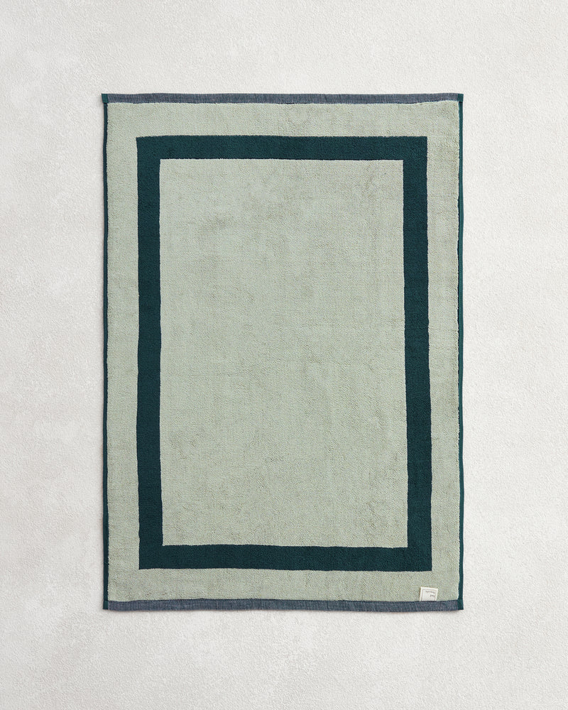 The Bed Threads Forest & Sage Two-Toned 100% Cotton Terry Bath Mat, in light green with a dark green double border and a small white label in one corner, rests on a textured surface.