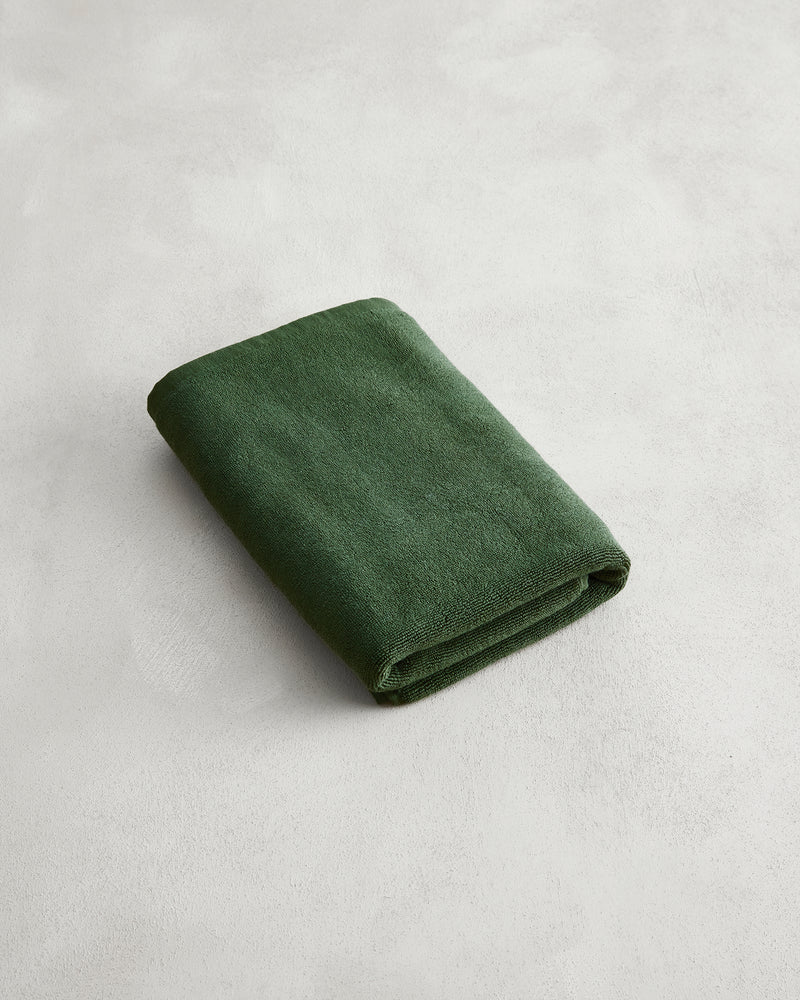 A neatly folded Moss 100% Cotton Terry Bath Mat by Bed Threads in dark green rests on a light gray textured surface, adding comfort and style.