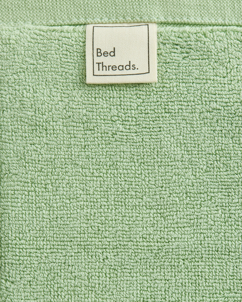 A close-up of the Apple 100% Cotton Terry Bath Mat by Bed Threads shows its light green texture and a white label at the top center, highlighting plush comfort and absorbency.