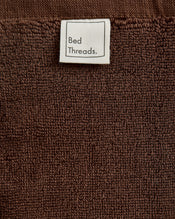 A close-up of the Espresso 100% Cotton Terry Bath Mat by Bed Threads, featuring a brown textured surface and a white label at the top with Bed Threads in black text. - view