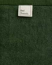 A close-up of Moss 100% Cotton Terry Face Washers (Set of Two) by Bed Threads, featuring dark green absorbent terry fabric and a white Bed Threads label—perfect for your daily skincare routine. - view