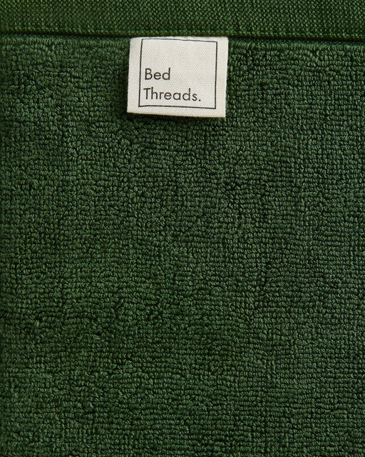 A close-up of Moss 100% Cotton Terry Face Washers (Set of Two) by Bed Threads, featuring dark green absorbent terry fabric and a white Bed Threads label—perfect for your daily skincare routine.