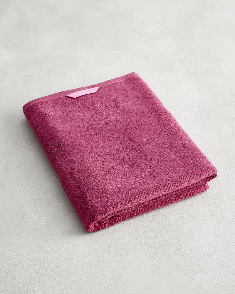 The Bed Threads Berry 100% Cotton Terry Bath Sheet, in plush raspberry, is neatly folded on a light gray surface with a light pink hanging loop visible. Quick-drying and stylishly functional, it's the perfect towel for any bathroom.