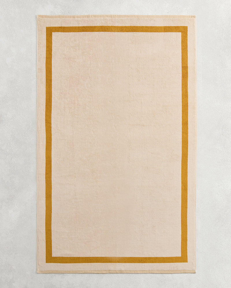 The Bed Threads Crème & Turmeric Two-Toned 100% Cotton Terry Bath Sheet adds cozy style to your bathroom, especially when paired with a rectangular beige rug on a light grey textured surface for an inviting touch.