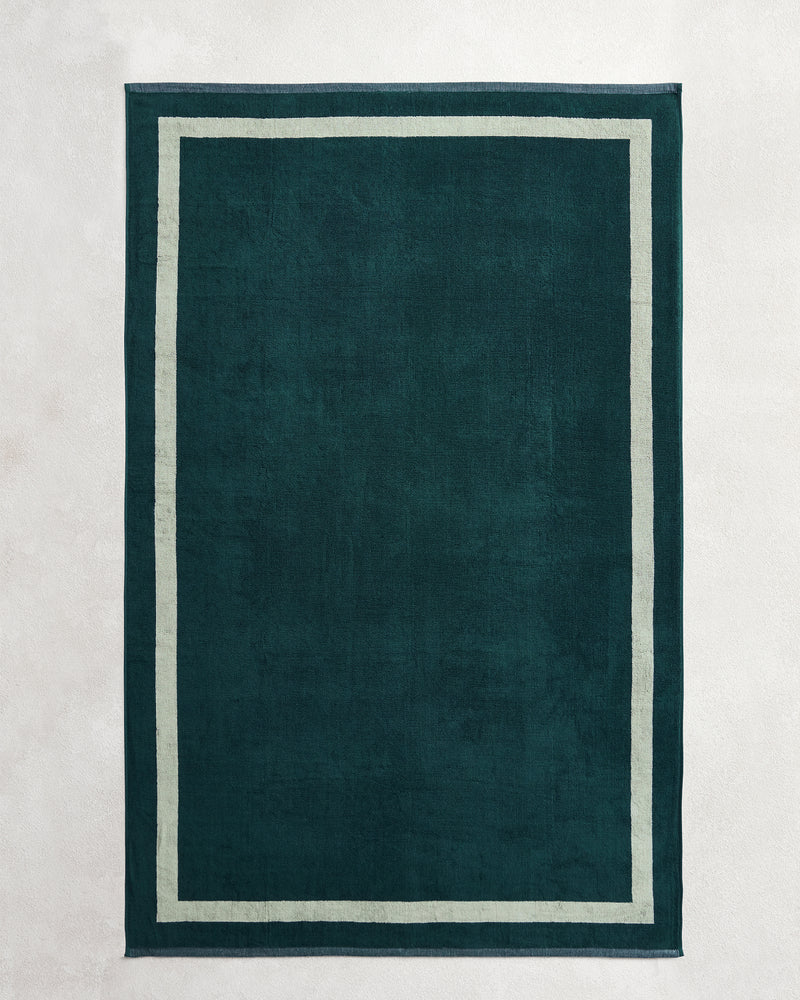 A dark green rectangular rug with a light border, inspired by the elegant Forest & Sage Two-Toned 100% Cotton Terry Bath Sheet Bundle from Bed Threads, lies on a pale, textured surface.