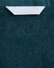 The Aegean 100% Cotton Terry Bath Sheet by Bed Threads features a plush, textured teal fabric with a rectangular white label at the top center, partially tucked under the edge. - view