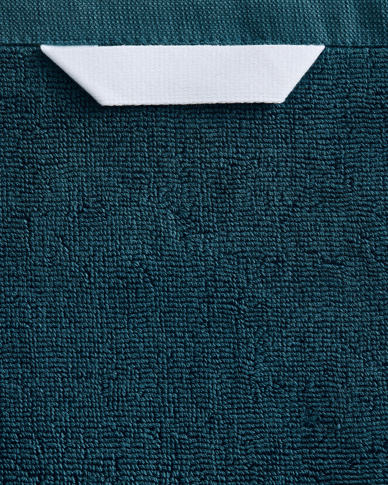 The Aegean 100% Cotton Terry Bath Sheet by Bed Threads features a plush, textured teal fabric with a rectangular white label at the top center, partially tucked under the edge.