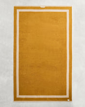 The Bed Threads Crème & Turmeric Two-Toned 100% Cotton Terry Bath Sheet, featuring a golden-yellow and white design, is shown laid flat on a light gray textured surface. - view