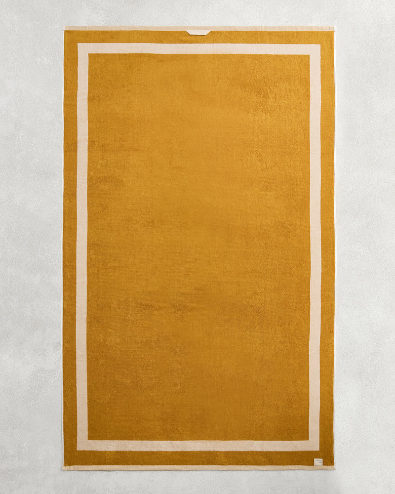 The Bed Threads Crème & Turmeric Two-Toned 100% Cotton Terry Bath Sheet, featuring a golden-yellow and white design, is shown laid flat on a light gray textured surface.
