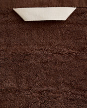 A close-up of the Bed Threads Espresso 100% Cotton Terry Bath Sheet shows its plush brown texture and a small white rectangular tag partially tucked beneath the top edge. - view