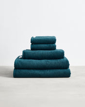 A neatly stacked set of four Aegean 100% Cotton Terry Bath Sheet Bundle towels by Bed Threads, in dark teal and varied sizes, arranged largest to smallest on a textured surface against a plain white background. - view