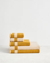 The Bed Threads Crème & Turmeric Two-Toned 100% Cotton Terry Bath Sheet Bundle features four luxury towels—two with wide beige stripes—neatly stacked on a light surface against a plain white wall. - view