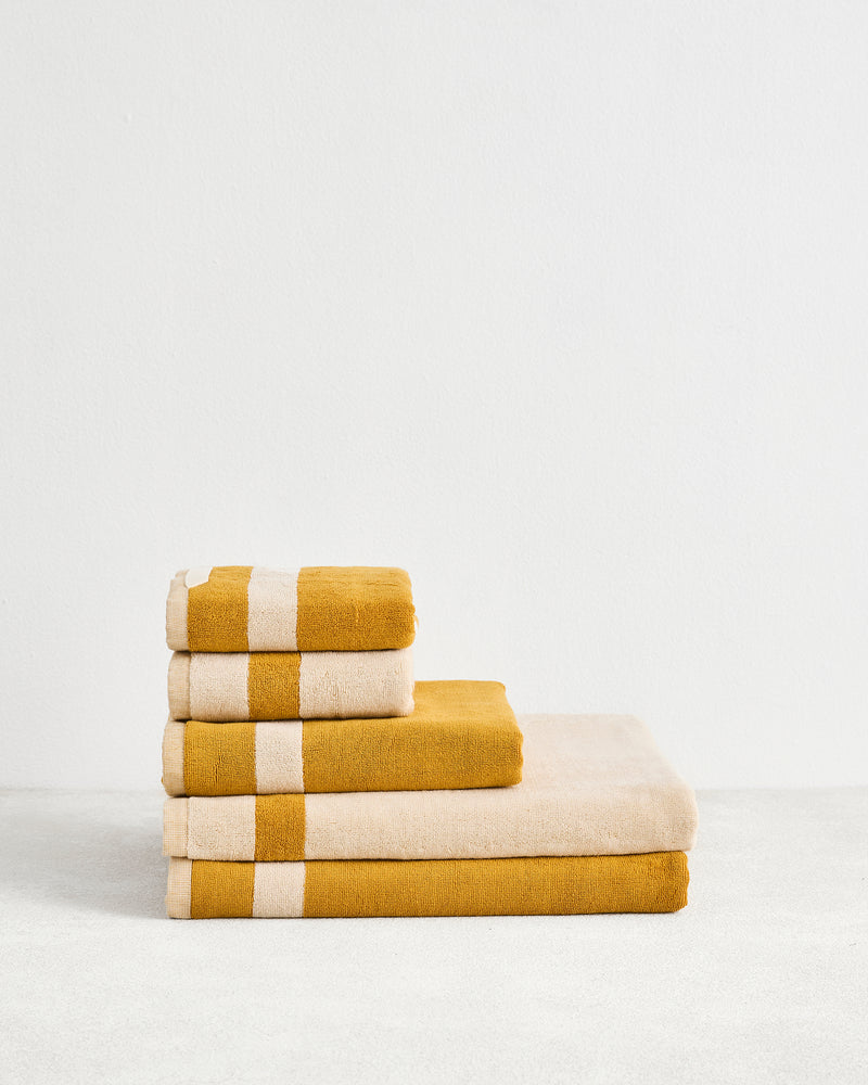 The Bed Threads Crème & Turmeric Two-Toned 100% Cotton Terry Bath Sheet Bundle features four luxury towels—two with wide beige stripes—neatly stacked on a light surface against a plain white wall.