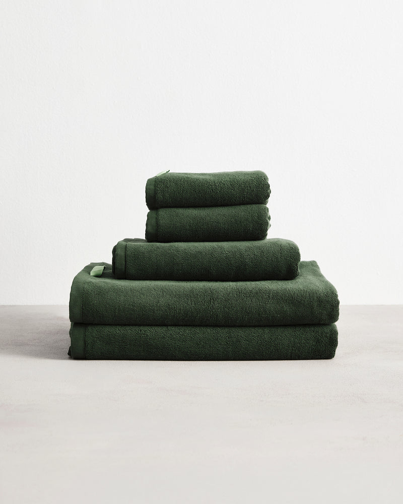 The Bed Threads Moss 100% Cotton Terry Bath Sheet Bundle, featuring bath sheets, towels, and hand towels in dark green, is neatly stacked on a light surface against a plain white background.