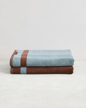 Two Cacao & Lagoon Two-Toned 100% Cotton Terry Bath Sheets by Bed Threads are neatly folded on a light surface, one light blue with a brown stripe atop a brown towel with a blue stripe. A plain white background highlights these luxury bath sheets. - view