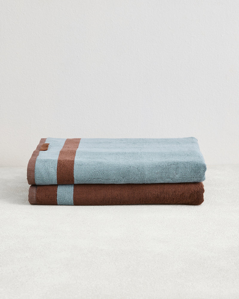 Two Cacao & Lagoon Two-Toned 100% Cotton Terry Bath Sheets by Bed Threads are neatly folded on a light surface, one light blue with a brown stripe atop a brown towel with a blue stripe. A plain white background highlights these luxury bath sheets.